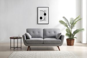 A contemporary Oslo 2 Seater Sofa in a textured grey fabric, featuring a clean-lined silhouette with tufted back cushions and two matching side pillows. The sofa is supported by angled wooden legs, giving it a mid-century modern feel. Positioned in a minimalist setting, the sofa is flanked by a round side table with books and a glass, and a lush potted palm to the right, enhancing the fresh, modern aesthetic. Above the sofa hangs a framed abstract monochrome artwork, against a pristine white wall. The setting is completed with a cream textured rug on a light wooden floor, creating a serene and inviting space.