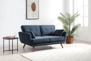 A contemporary Oslo 2 Seater Sofa in a textured blue ink fabric, featuring a clean-lined silhouette with tufted back cushions and two matching side pillows. The sofa is supported by angled wooden legs, giving it a mid-century modern feel. Positioned in a minimalist setting, the sofa is flanked by a round side table with books and a glass, and a lush potted palm to the right, enhancing the fresh, modern aesthetic. Above the sofa hangs a framed abstract monochrome artwork, against a pristine white wall. The setting is completed with a cream textured rug on a light wooden floor, creating a serene and inviting space.