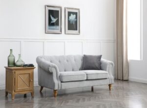 Elegant three-seater Chesterfield sofa in a light grey fabric, featuring a classic low back style with deep buttoned arms and back, complemented by a tufted front border. The sofa's scroll fronted arms are adorned with lines of individual stud nails and supported by turned wooden legs. It is placed in a bright, well-lit room with minimalist decor, including a wooden side table, a couple of green vases, and two framed botanical prints on the wall behind it. The room's aesthetic is completed with a light-coloured wooden floor and long, flowing curtains on the window to the right