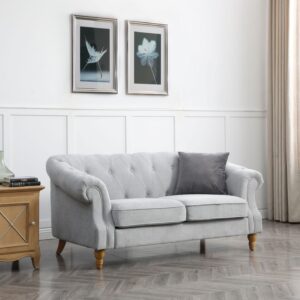 Elegant three-seater Chesterfield sofa in a light grey fabric, featuring a classic low back style with deep buttoned arms and back, complemented by a tufted front border. The sofa's scroll fronted arms are adorned with lines of individual stud nails and supported by turned wooden legs. It is placed in a bright, well-lit room with minimalist decor, including a wooden side table, a couple of green vases, and two framed botanical prints on the wall behind it. The room's aesthetic is completed with a light-coloured wooden floor and long, flowing curtains on the window to the right