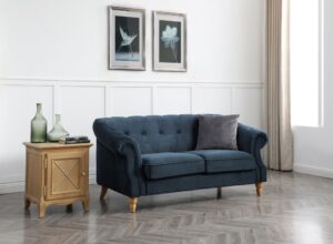 A luxurious ink velvet three-seater Chesterfield sofa with a classic silhouette, featuring a tufted back and arms with deep button detailing. The sofa's rolled arms are highlighted with nailhead trim, presenting a sophisticated aesthetic. It stands on elegantly turned wooden legs with a natural finish, contrasting beautifully with the dark, rich velvet. To its left is a wooden side table with an X-patterned cabinet door, upon which rest two green glass vases and a stack of books. The backdrop consists of a white paneled wall adorned with two framed botanical prints, with natural light streaming in from a curtained window to the right, completing the serene and stately scene.