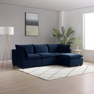 A luxurious Kyoto Moda corner sofa in sumptuous navy velvet, featuring a chaise lounge for added comfort. This elegant piece is the centerpiece of a modern living room with light wooden flooring and grey walls. A stylish tripod floor lamp with a white shade and a textured white and black area rug complement the deep blue of the sofa. Natural light streams in from the window, highlighting the lush green potted plant and a side table with delicate flowers, creating an ambiance of sophisticated comfort.