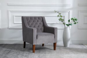 A sophisticated grey Isabel Chesterfield chair by Kyoto, featuring classic deep button tufting on the backrest and armrests. The chair is set in an elegant room with white panelled walls, standing on dark wooden legs. To its right, a tall white vase holds a sprig of green eucalyptus leaves, adding a touch of natural beauty. The scene is completed with a soft grey area rug beneath the chair, contributing to the room’s refined and serene atmosphere.
