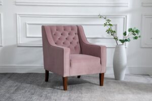 An inviting Isabel Chesterfield armchair in heather pink velvet, with classic deep button detailing on the backrest, complemented by dark wooden legs. Positioned on a grey rug against a white panelled wall, the chair is accented with a tall white textured vase filled with eucalyptus branches to its side, enhancing the chair's elegant appeal and the room's overall sense of calm sophistication.