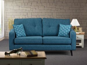 A Wentworth sofa in a vibrant Aegean blue colour, featuring a robust frame with a textured fabric finish. The sofa has two plush seat cushions and is accented with two geometric-patterned throw pillows. It is set against a room with a stone-brick wall painted in muted grey, beside a white side table with a vintage radio and lamp, and a model airplane and book on a coffee table in the foreground, creating a cosy and stylish living space.