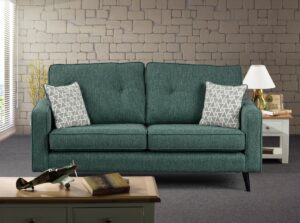 A comfortable Wentworth sofa in a rich green fabric, with two matching back cushions and a geometric-patterned throw pillow, set in a contemporary living room. The sofa stands on sleek black legs against a muted stone brick wall, alongside a classic white side table with a vintage radio and a table lamp. In the foreground, a model airplane and a historical book rest on a wooden coffee table, adding character to the inviting scene.