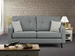 A chic Wentworth sofa in a sophisticated silver fabric, featuring clean lines and two plush seat cushions, accessorized with two white geometric-patterned throw pillows. The sofa is set against a modern room backdrop with a textured stone brick wall and soft yellow accents from a decorative branch. Completing the scene is a white side table with a classic lamp and vintage radio, and a model airplane alongside a book on a wooden coffee table in the foreground, blending contemporary style with nostalgic decor.