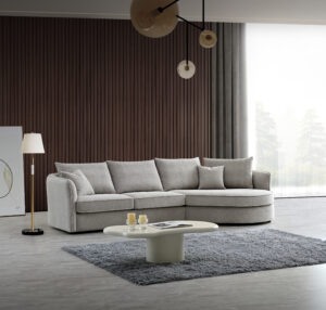 Modern and inviting living room featuring the Rubin Corner Sofa upholstered in Pebble Bouclé. The sofa is arranged in a spacious setting with a dark striped backdrop and large windows revealing a misty forest view. The room is complemented by a contemporary white coffee table, a plush grey area rug, and stylish lighting with a floor lamp and overhead bulb pendants.