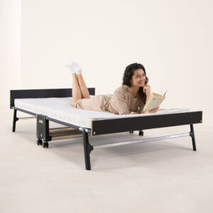 A woman lying comfortably on the Jay-Be GP120 Grand Folding Bed, reading a book. The bed features a sturdy black frame with a headboard and footboard, and a comfortable e-Pocket™ mattress.