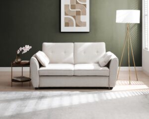 A cream-colored Kyoto Willow sofa bed placed in a bright, modern living room setting with a side table, lamp, and decorative art piece in the background.